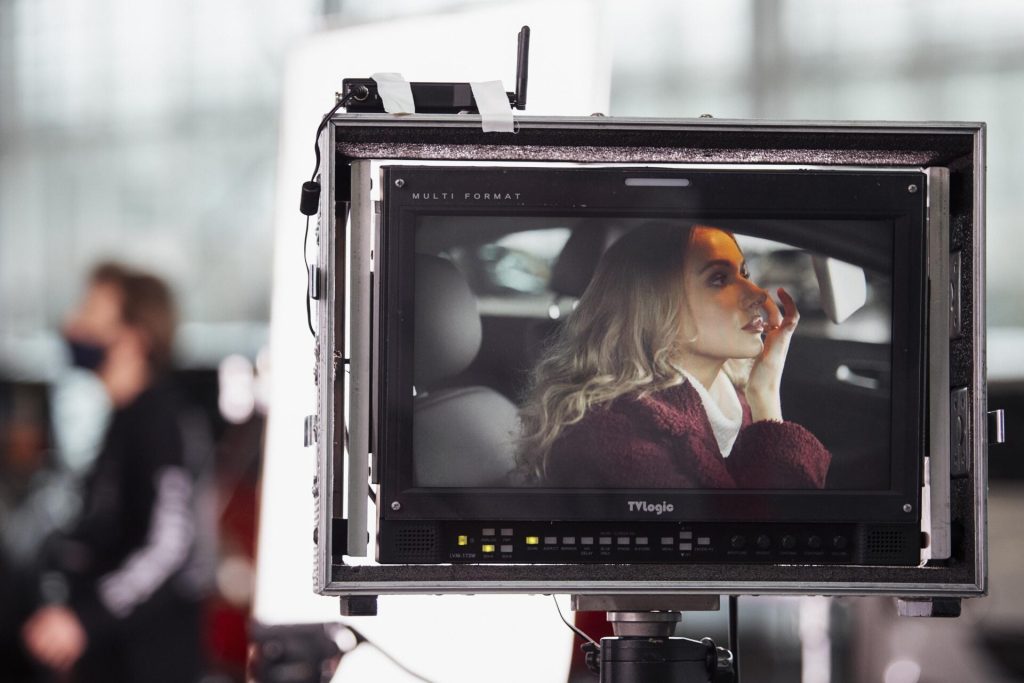 A shot of an actress through a directors monitor checking her make up in the rear view mirror of a car while filming an advertisement